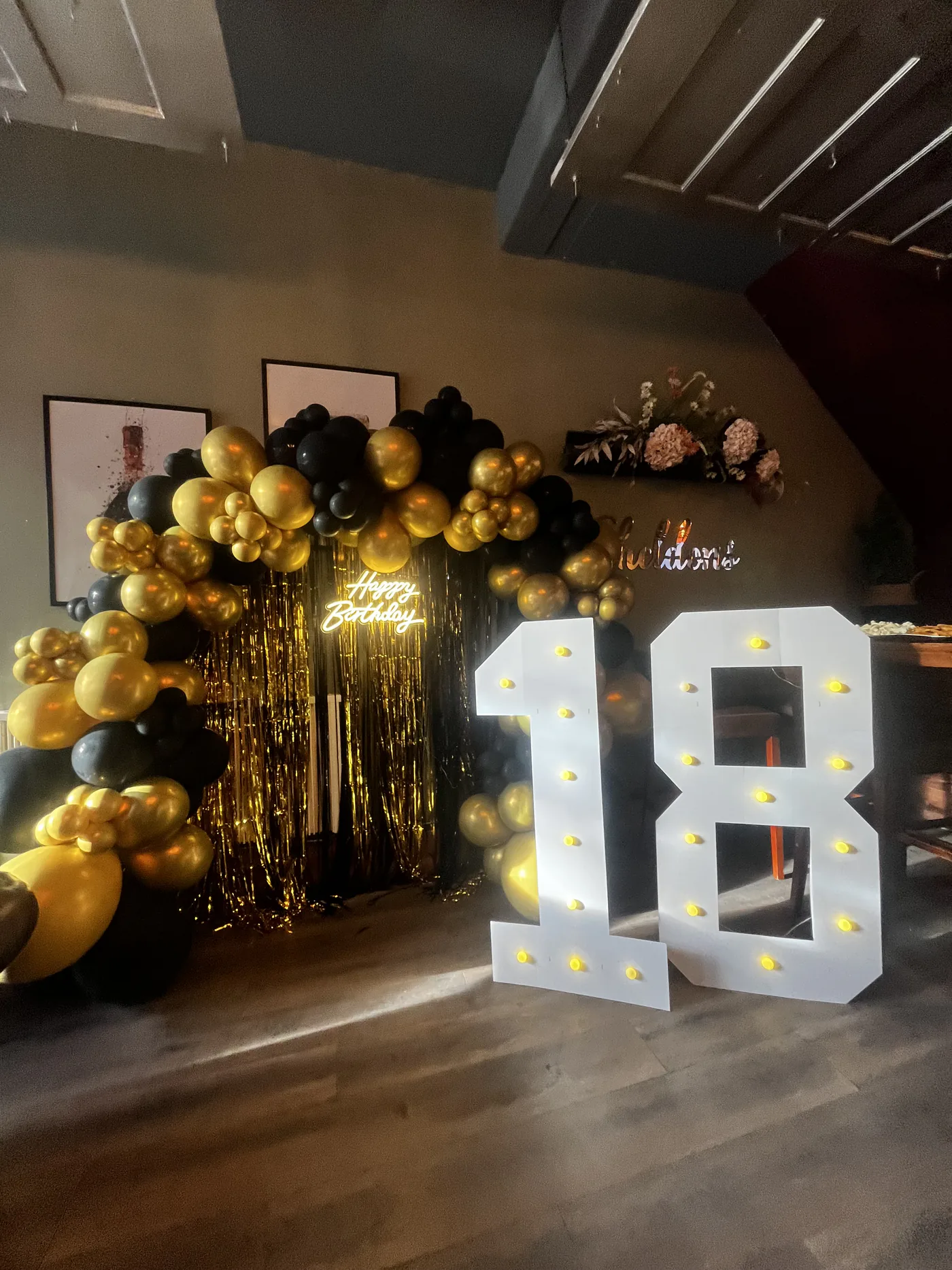 A milestone birthday setup with a black and gold balloon arch, a gold fringe backdrop, a neon happy birthday sign, and illuminated marquee number lights.