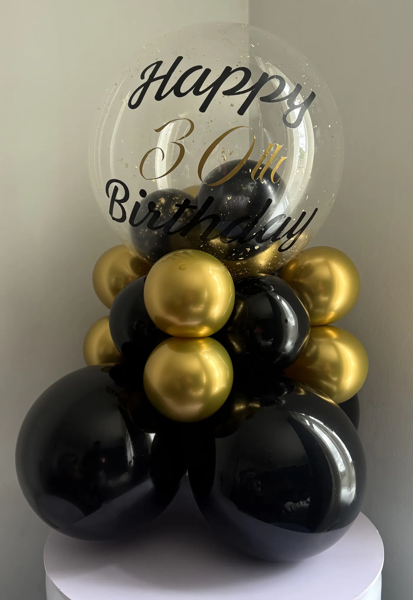 A milestone birthday balloon display in black and gold, topped with a printed clear bubble balloon and set on a white plinth.