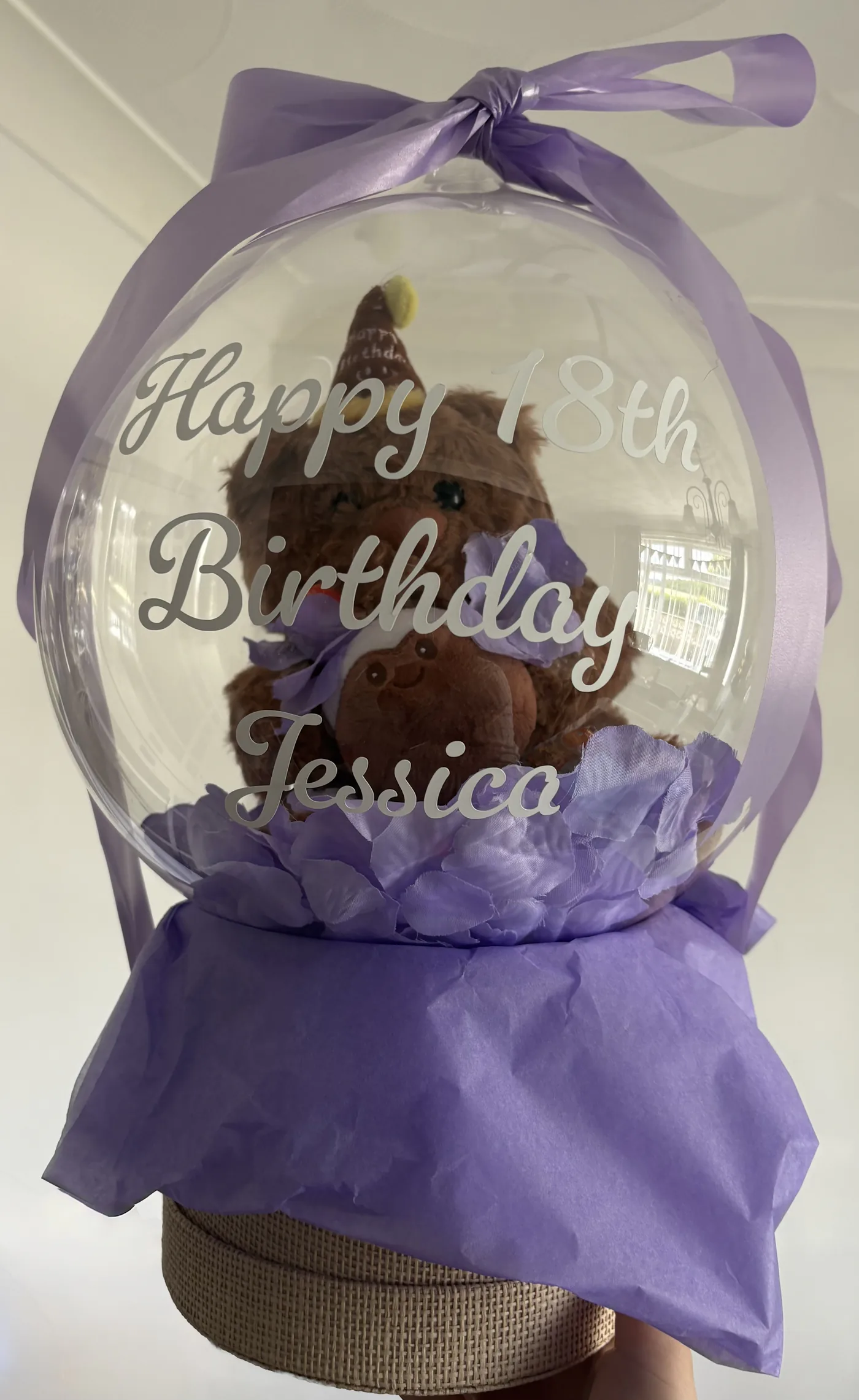 A personalised clear bubble balloon tied with ribbon, containing a soft toy and petals, presented on a woven gift base.