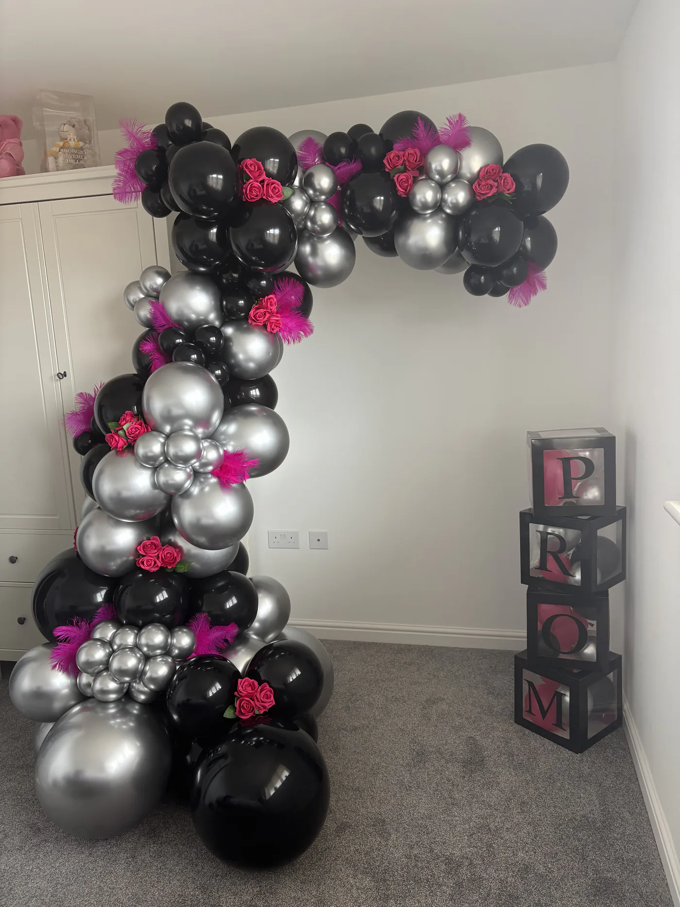 A hand-built balloon arch in black, chrome silver and hot pink, finished with roses and feathers, beside stacked lettered display boxes.